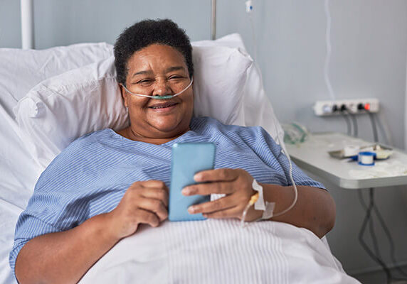 iStock-1469539615 WEB A smiling woman is in a hospital bed and is propped up by pillows. She has breathing tubes in her nose and an IV in her hand. She is using her cell phone to order food.